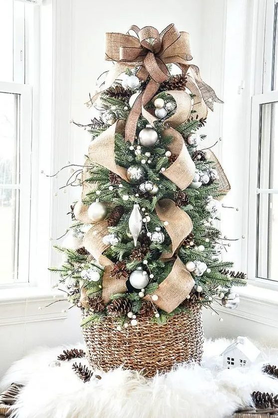 a gorgeous tabletop Christmas tree with silver and metallic ornaments, pinecones, twigs and a large burlap bow on top