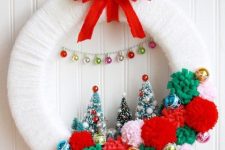 a white Christmas wreath with colorful pompoms, ornaments and bottle bursh Christmas trees, a red bow and ribbon