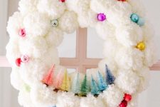 a white pompom Christmas wreath with bright and shiny ornaments and little tinsel trees of all the colors of rainbow