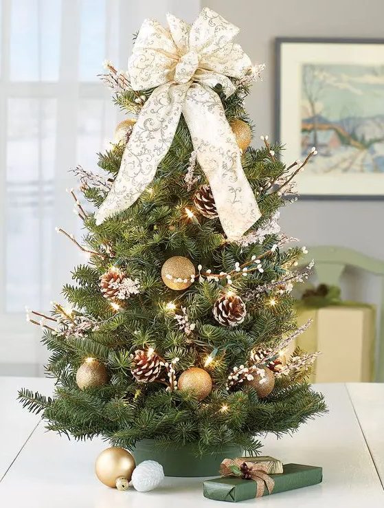 an elegant small Christmas tree with gold glitter ornaments, snowy pinecones, lights, willow and a large white and gold bow on top