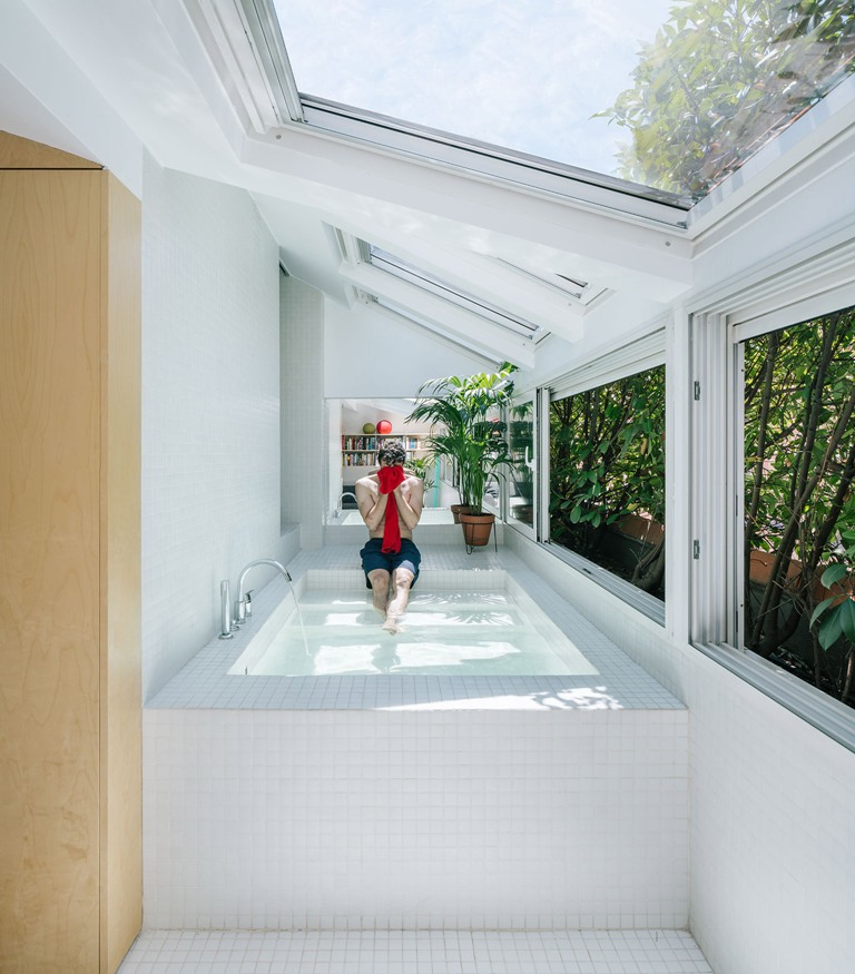 The bathtub is a large pool like space clad with white tiles, with skylights and windows that show off greenery walls