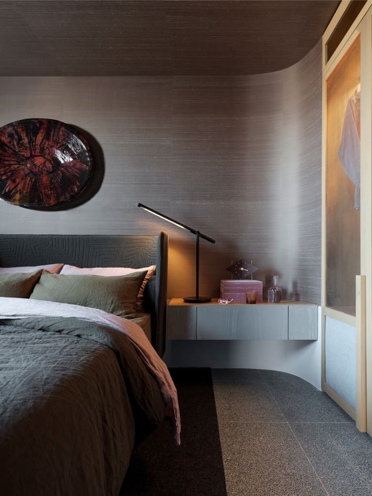 The bedroom features a beautiful rounded wall, an upholstered bed, artworks and floating bedside tables