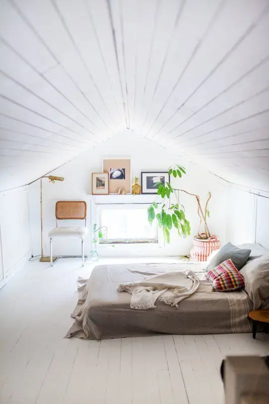 An all white attic bedroom with neutral wood and textiles, with potted greenery and artworks