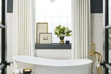 a luxorious bathroom with a black and white tile floor, white subway tiles and black walls, a chic tub, gold fixtures and a lovely chandelier