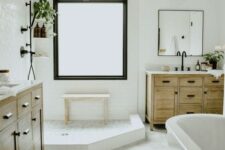 a neutral farmhouse bathroom with a two wooden vanities, an oval tub, a shower space with a large window with frosted glass