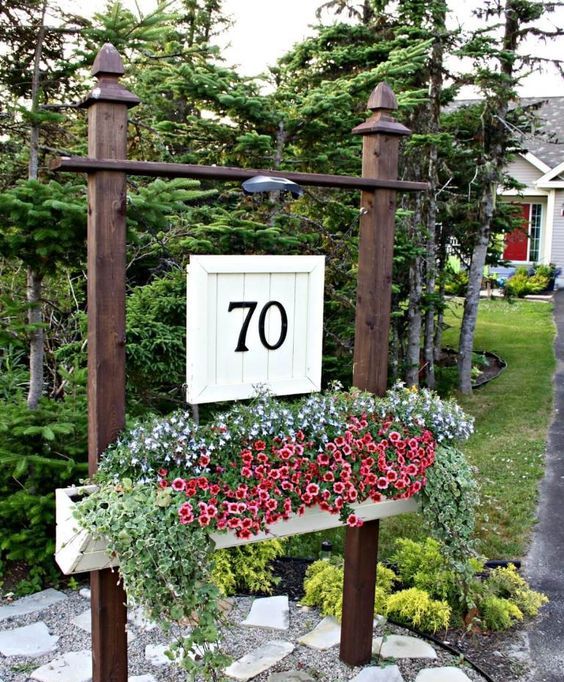 a large outdoor planter on stands with lush blooms and greenery and a wooden plaque with house numbers and an additional light