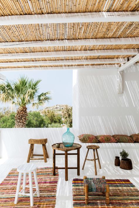 a tropical terrace with colorful boho rugs, wooden stools and a table, some pillows and potted grreenery