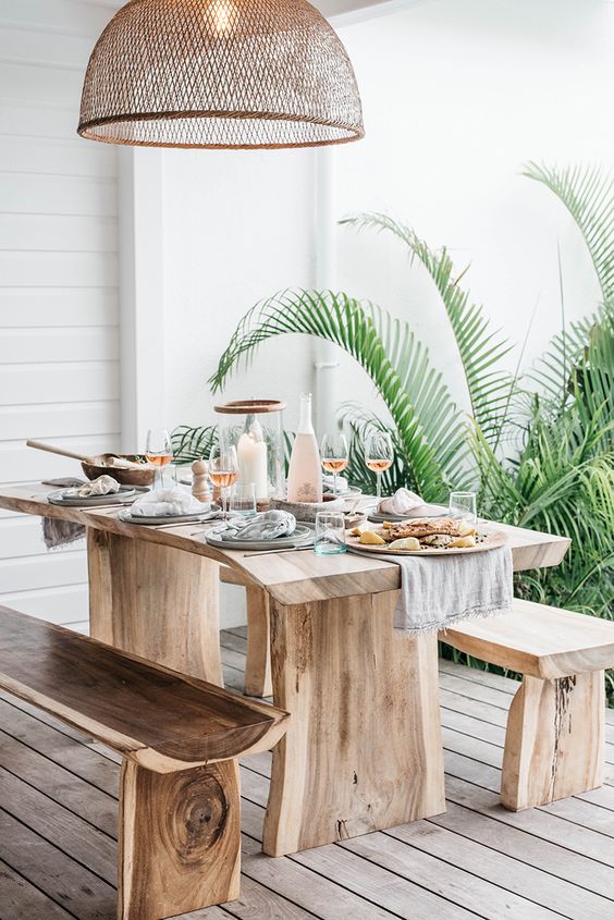 a tropical outdoor dining space with rough wooden furniture, a wicker lampshade and potted greenery