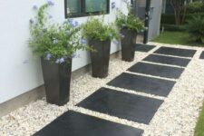 a stylish modern front yard with gravel and oversized black tiles, tall black planters with greenery and blooms