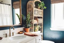 a moody eclectic bathroom with soot walls, a white vanity, a rattan shelving unit, white appliances and some potted greenery