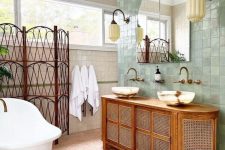 an eclectic bathroom clad with neutral and green tile, pink printed tile on the floor, a space divider, a cane vanity, a tub and some vintage lamps