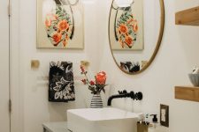 an eclectic bathroom with a light grey vanity, a round mirror, a bold artwork, wooden shelves and a sink with a black faucet