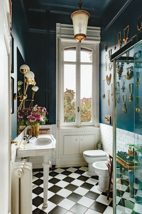 An eclectic bathroom with navy walls and a gallery wall of jewelry, a vintage free standing sink, glam lamps and gold accessories