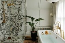 an eclectic bathroom with neutral paneling, a white marble shower space, a tub, a rug, a potted plant and a chandelier