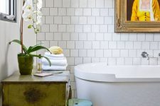 an eclectic bathroom with square and colorful tiles, an oval tub, a vintage vanity, a green acrylic stool and an artwork
