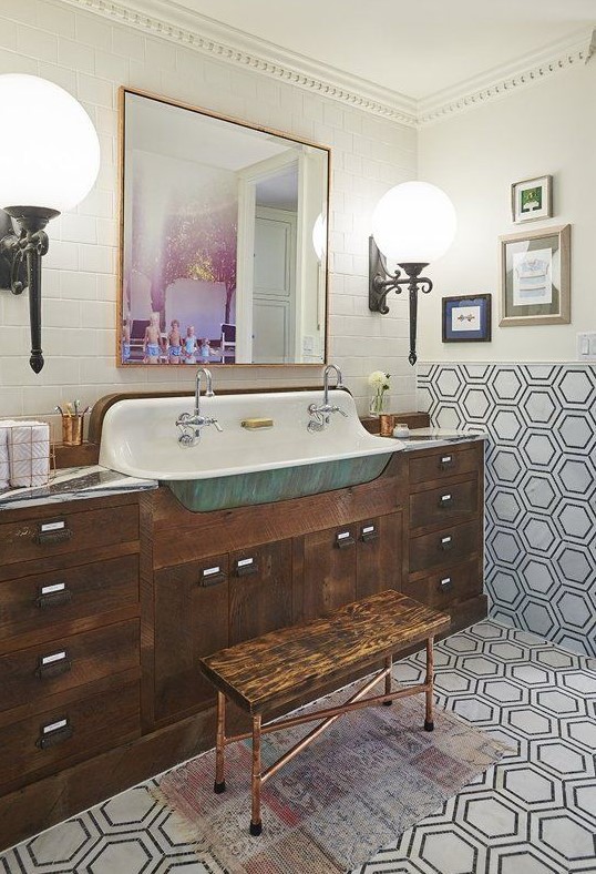 An eclectic bathroom with subway and hexagon tiles, a dark stained vanity, a stool, a gallery wall and large sconces