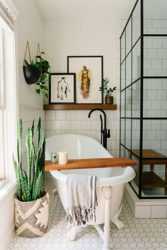 An eclectic bathroom with white square tiles, a free standing tub,  a shower space, a shelf with art and potted greenery