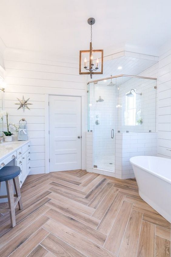 A light filled farmhouse bathroom featuring white shiplap, a wooden floor, a cool chandelier and a free standing tub