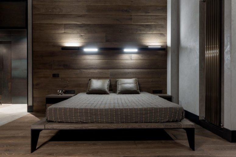 The bedroom shows off a wooden accent wall, a wooden bed with legs and some lights over the bed