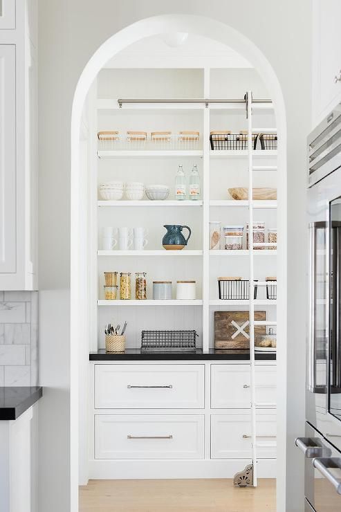 an arched doorway into a custom pantry featuring a ladder on rails on a built-in shelf with black quartz countertops