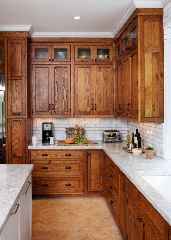 A rustic kitchen with warm stained cabinets, white skinny tiles, white stoen countertops plus vintage handles
