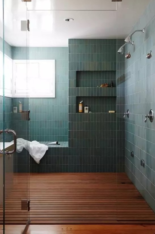 A chic bathroom with a large shower space clad with green stacked tiles, with a bench and niches in the walls, a dark stained floor and windows for natural light