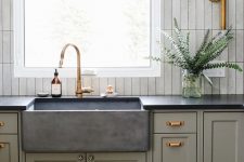 a chic kitchen with grey green cabinets, a stone sink and black countertops, a white skinny tile backsplash and brass touches