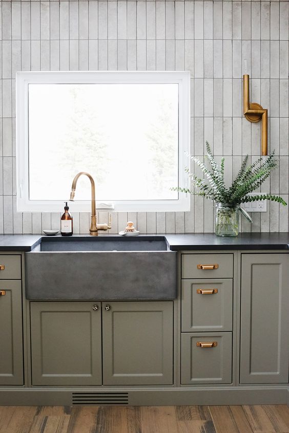 a chic kitchen with grey green cabinets, a stone sink and black countertops, a white skinny tile backsplash and brass touches