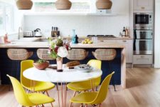 a cool tropical kitchen with a navy kitchen island plus white cabinets, rattan stools and wicker lamps, bright yellow chairs and a round table