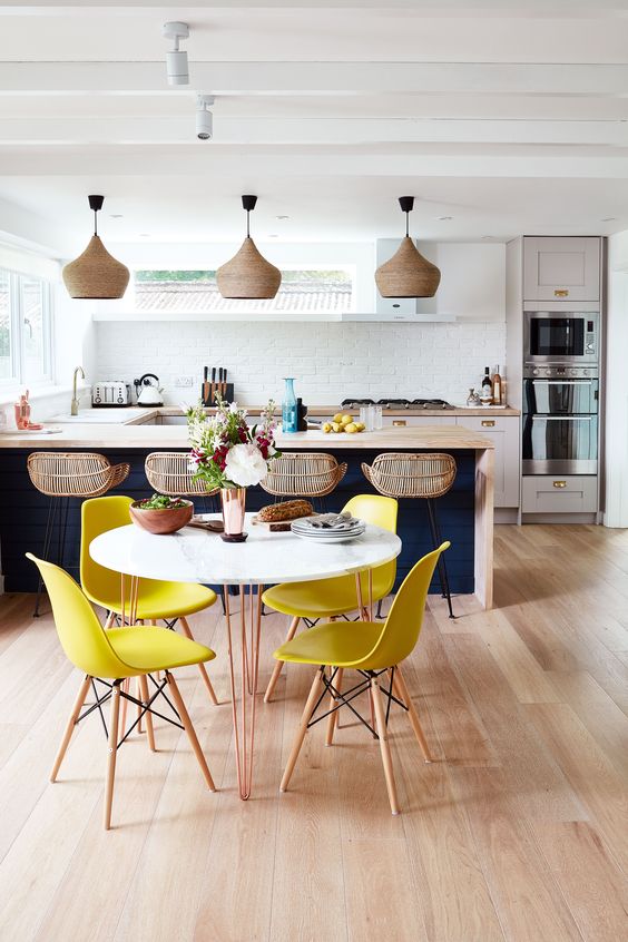 a cool tropical kitchen with a navy kitchen island plus white cabinets, rattan stools and wicker lamps, bright yellow chairs and a round table