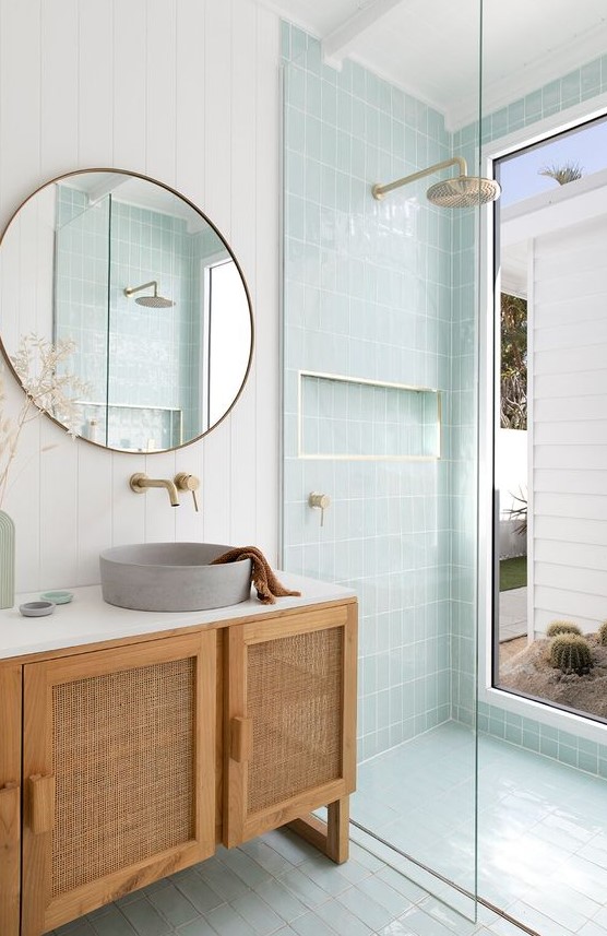 A dreamy mid century modern bathroom with white and mint blue tiles, a rattan vanity, a round mirror and a round sink