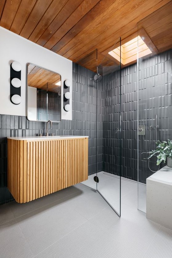 A mid century modern bathroom with graphite grey tiles, a fluted floating vanity, a shower space with a skylight