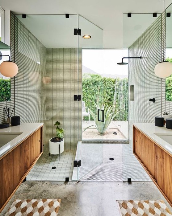 A mid century modern bathroom with skinny green tiles in the shower and a window, stained vanities, printed rugs and potted plants