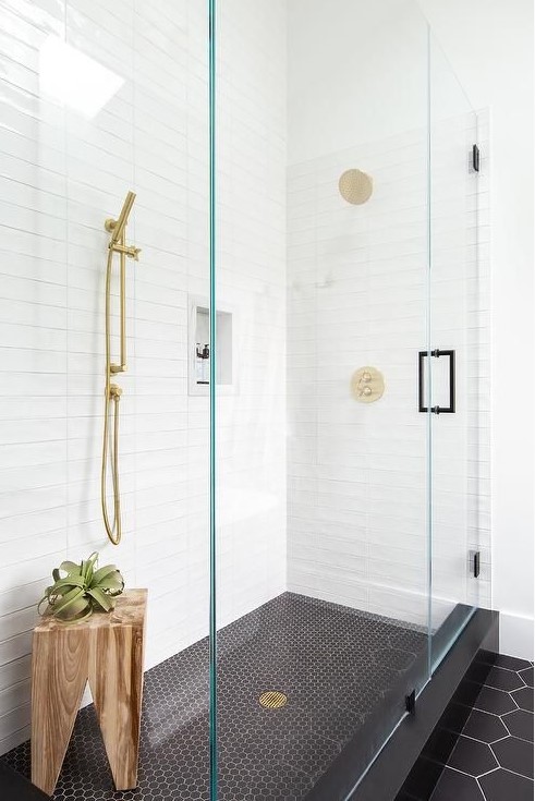 A mid century modern bathroom with stacked white tiles, black penny and hexagon ones, a wooden stool and brass touches