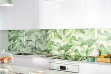 a modern tropical kitchen with sleek white cabinets, a tropical leaf print backsplash, green pendant lamps and wooden chairs