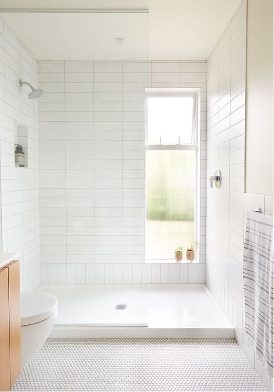 A neutral bathroom with stacked white tiles in the shower, penny tiles on the floor, a light stained floating vanity and a window