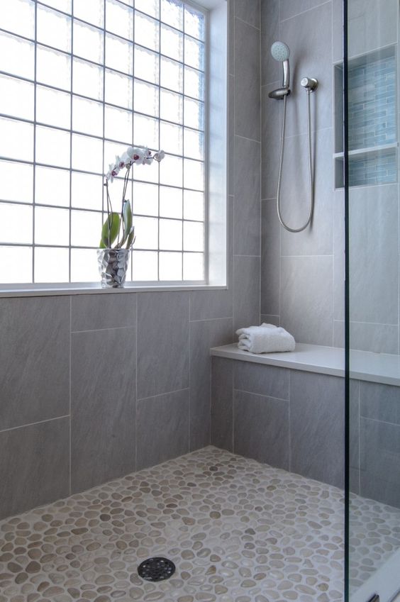 a neutral shower space with a pebble floor and grey tiles, a window with framing and a built-in bench
