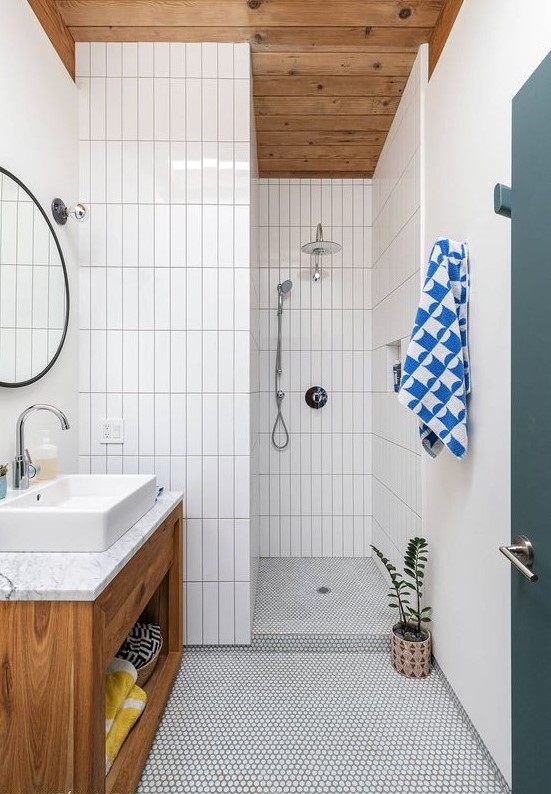 A simple mid century modern bathroom with white penny and skinny tiles, a wooden vanity and ceiling and a round mirror