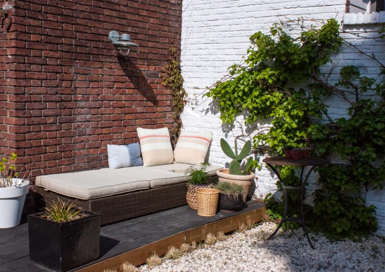 a small dark deck with an upholstered bench, potted greenery, cacti and succulents, a wall lamp and a side table