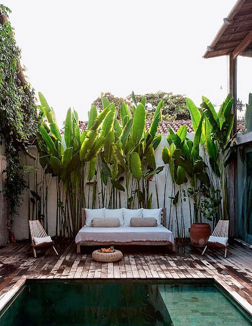a small deck with a comfortable daybed, some folding chairs, a jute ottoman and greenery
