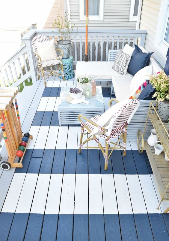 a small striped deck with rattan furniture, an L-shaped bench with pillows, potted greenery and a blue coffee table