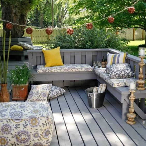 a small weathered deck with a built-in L-shaped bench, printed cushions and pillows, some potted greenery