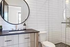 a stylish modern bathroom with white and black skinny tiles clad stacked, a timber vanity, a round mirror and a sconce