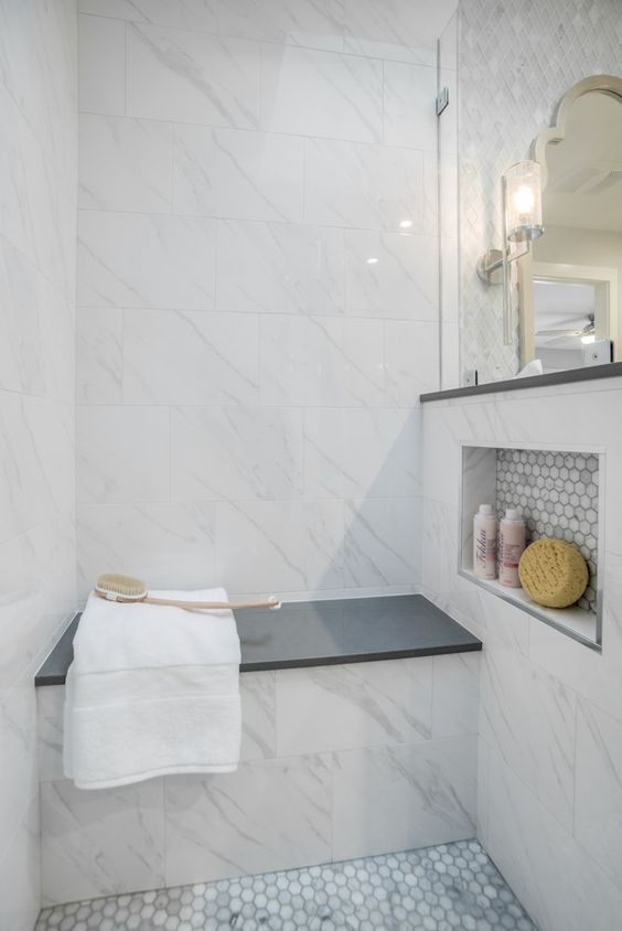 a stylish small shower space done with marble tiles, marble penny tiles, a built-in bench with a black seat
