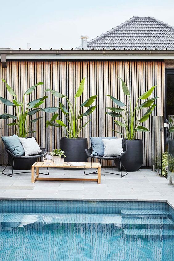 a tiled pool deck with a wooden table, some rattan chairs with pillows and potted greenery in black planters