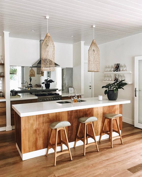 a tropical kitchen with white cabinets, stained wood covering the kitchen island, wooden stools, wicker lamps and a mirror backsplash
