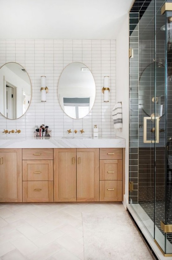 A two tone bathroom with white and black stacked tiles, a light stained vanity, oval mirrors, brass fixtures and sconces