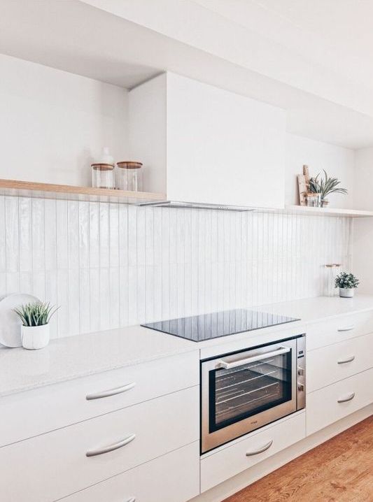 A white kitchen with sleek cabinets, skinny tiles, open shelves, a hood and built in appliances plus potted plants
