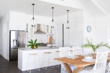 a white tropical kitchen with a marble backsplash, glass pendant lamps, wood and metal stools and a dining space with a wooden table and white rattan chairs