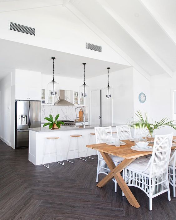 a white tropical kitchen with a marble backsplash, glass pendant lamps, wood and metal stools and a dining space with a wooden table and white rattan chairs